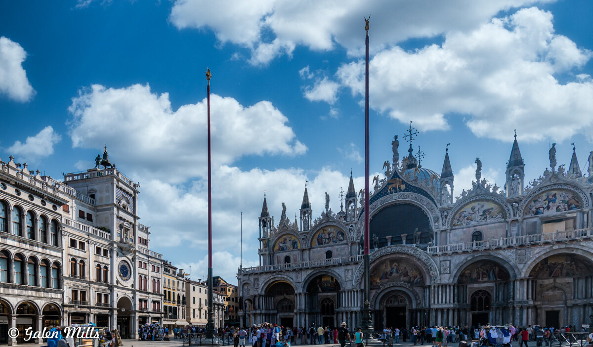 St. Mark's Basilica in Venice, Italy, with a clear blue sky and scattered clouds. It showcases the ornate architectural details and spires. Tourists are gathered in the foreground.