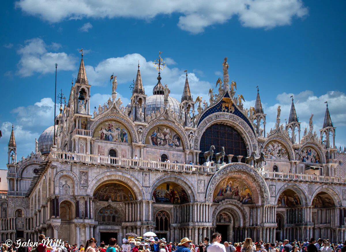 Front view of St. Mark's Basilica in Venice with tourists gathered in front, blue sky with clouds.