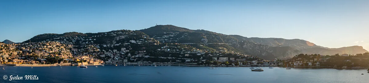Scenic coastal view with mountains, buildings, and yachts on the water at sunset.