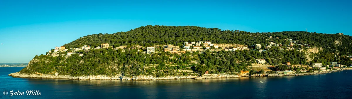 Scenic coastal landscape with a green, tree-covered hill dotted with houses, surrounded by calm, blue ocean waters under a clear sky.