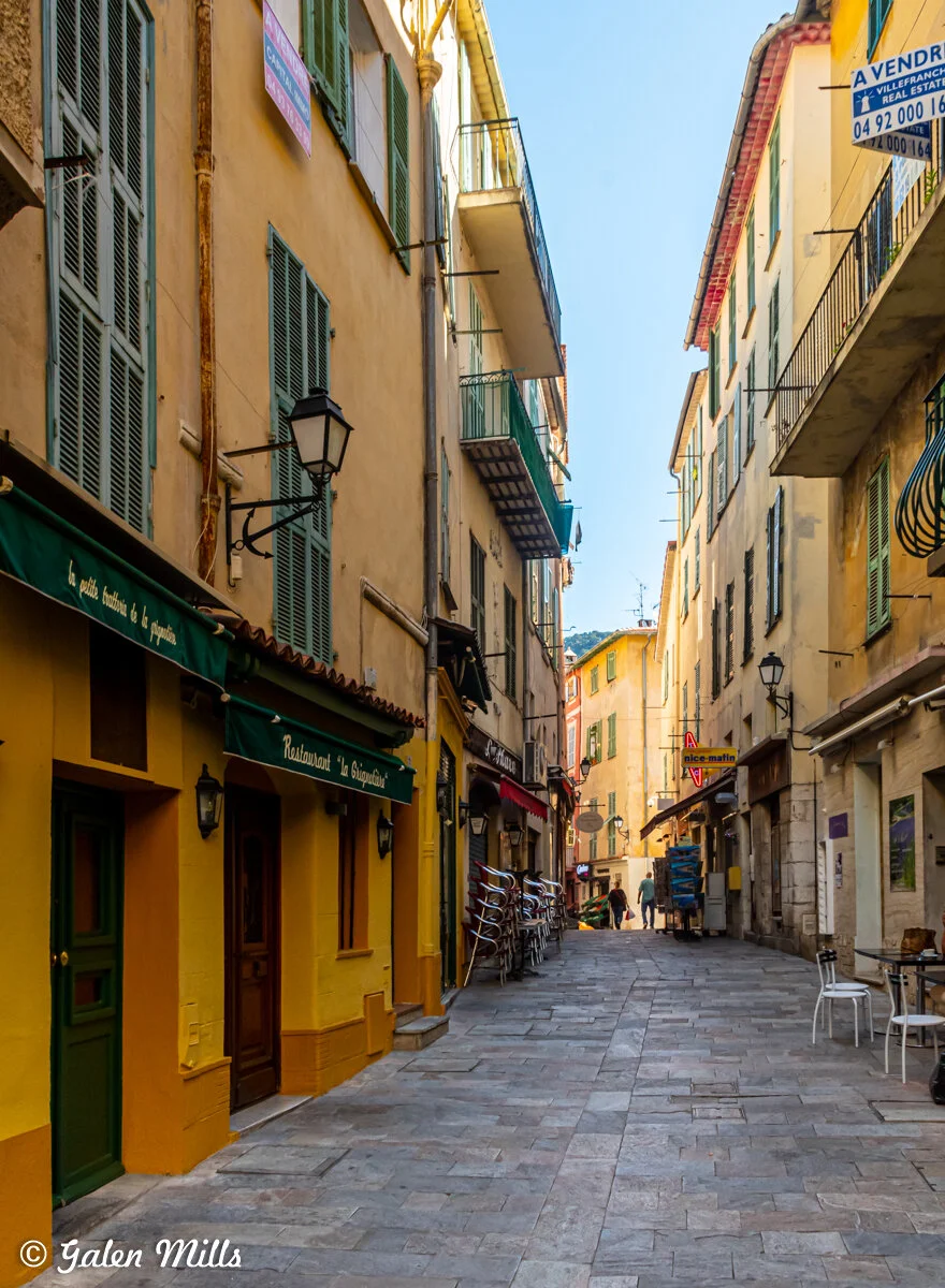 Narrow European street with colorful buildings, shutters, balconies, and hanging signs