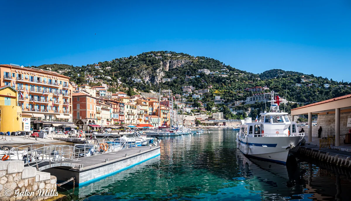 Port of Villefranche-sur-Mer with colorful buildings and boats, set against a hillside with greenery under a clear blue sky.
