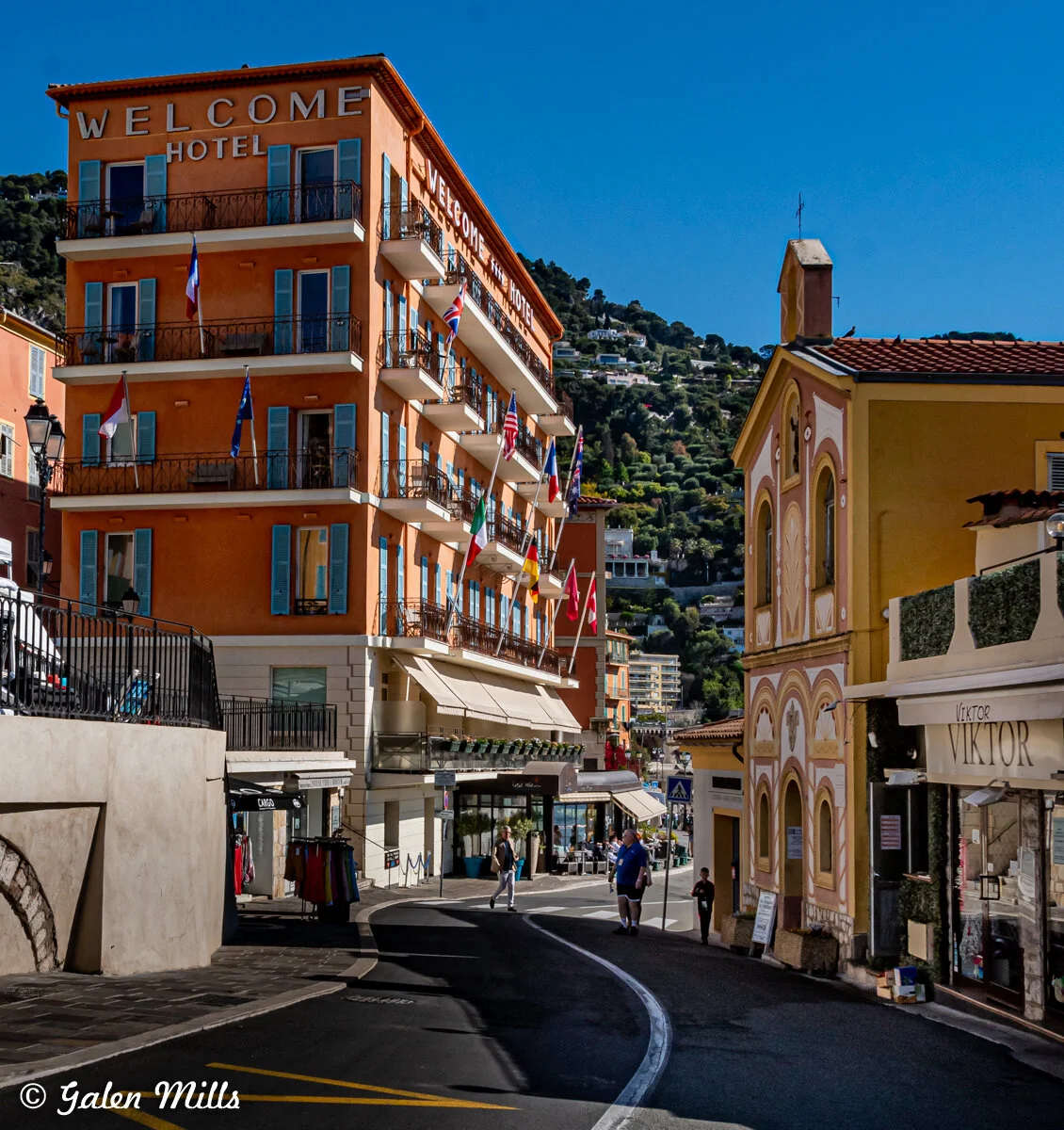 Street view in Villefranche-sur-Mer featuring Welcome Hotel and surrounding buildings with flags.
