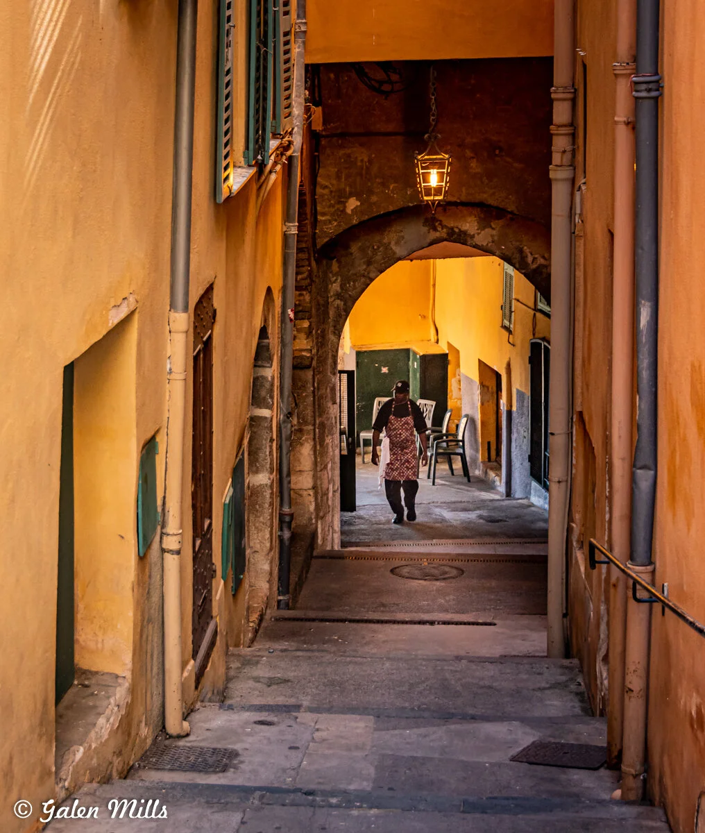 Narrow alleyway with yellow walls and a person walking, wearing an apron and cap, under an archway with a hanging lantern. Chairs are visible in the background.