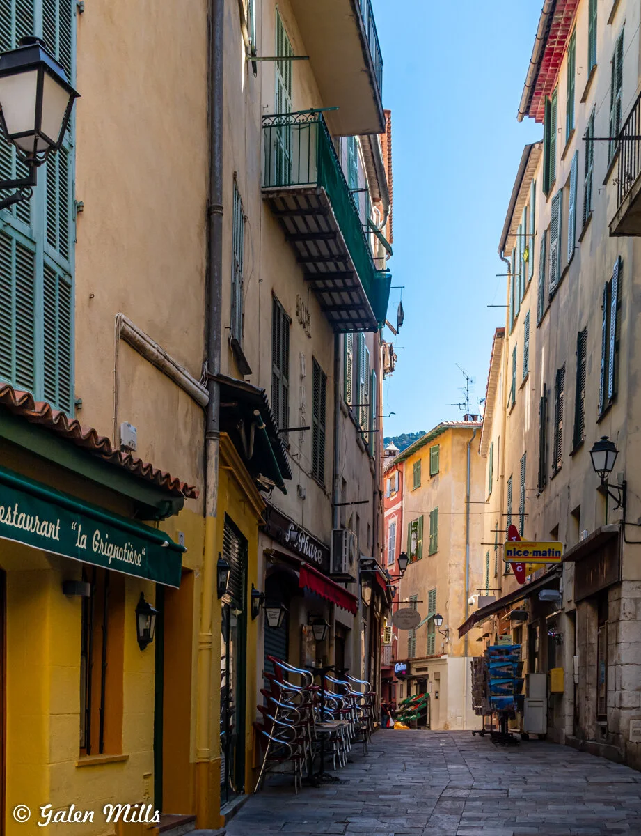 A narrow European street lined with tall, colorful buildings. The street features stacked chairs outside a café, street lamps, and hanging signs. The sky is clear and blue, adding a bright contrast to the shuttered windows and flower boxes.
