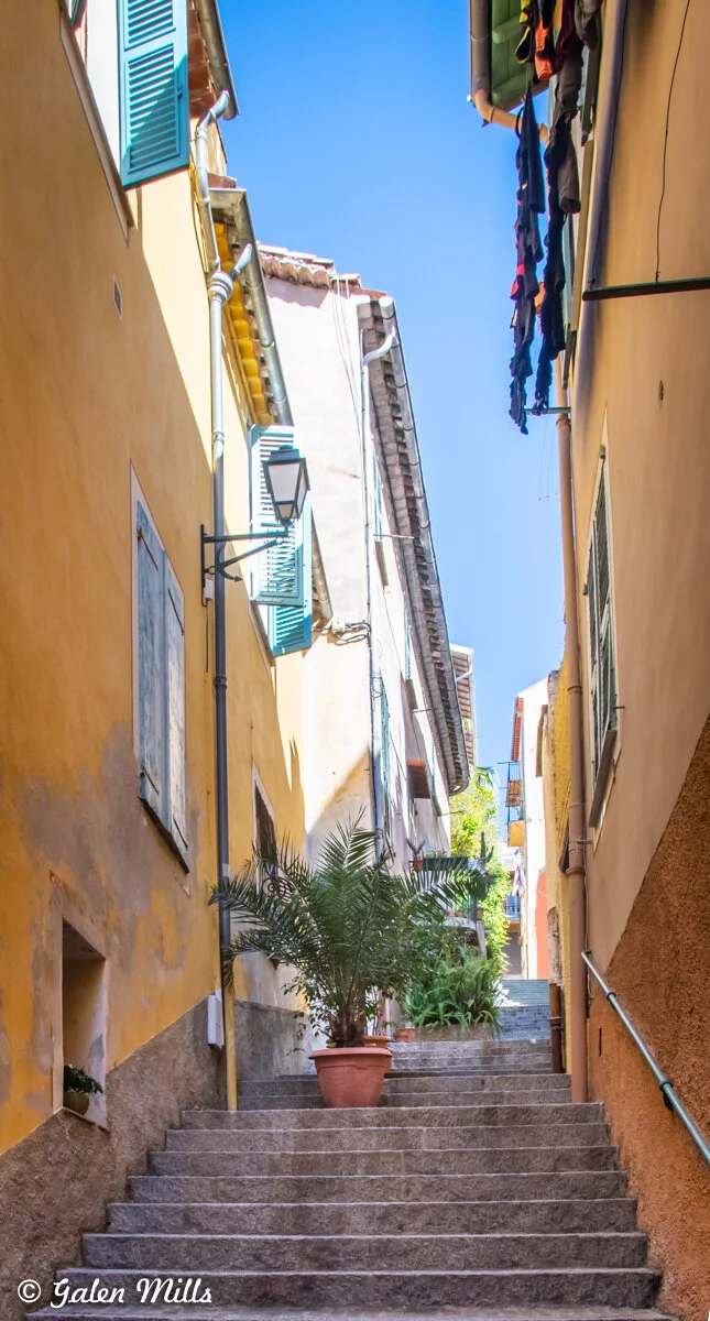 Narrow European alley with stone stairs, lined with colorful buildings and shutters, a potted plant, and drying clothes overhead.