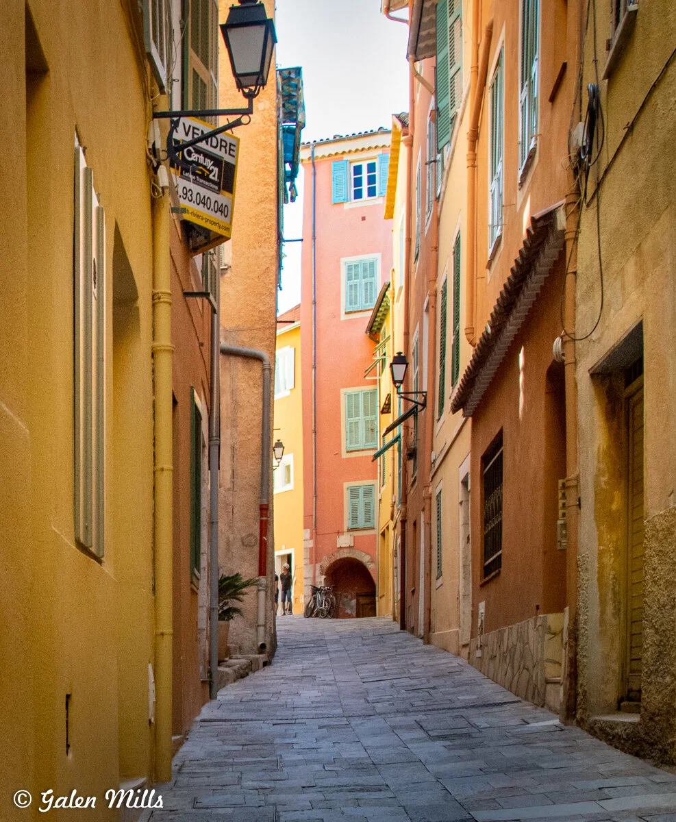 Narrow cobblestone alleyway with colorful European-style buildings, green shutters, and street lamps. A 'for sale' sign and bicycles are visible. Two people stand in the distance.