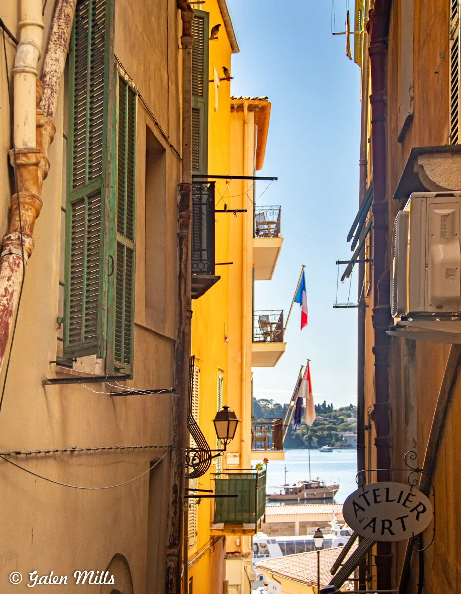Narrow alleyway in a coastal town with colorful buildings, balconies, and French flags. The image shows a view towards the sea and a boat in the distance under a clear blue sky. A sign for "Atelier d'Art" is visible.