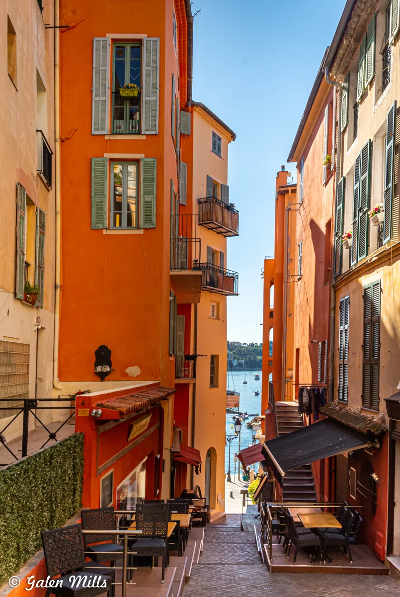 Narrow European street with colorful buildings, outdoor seating, and view of boats on the water.