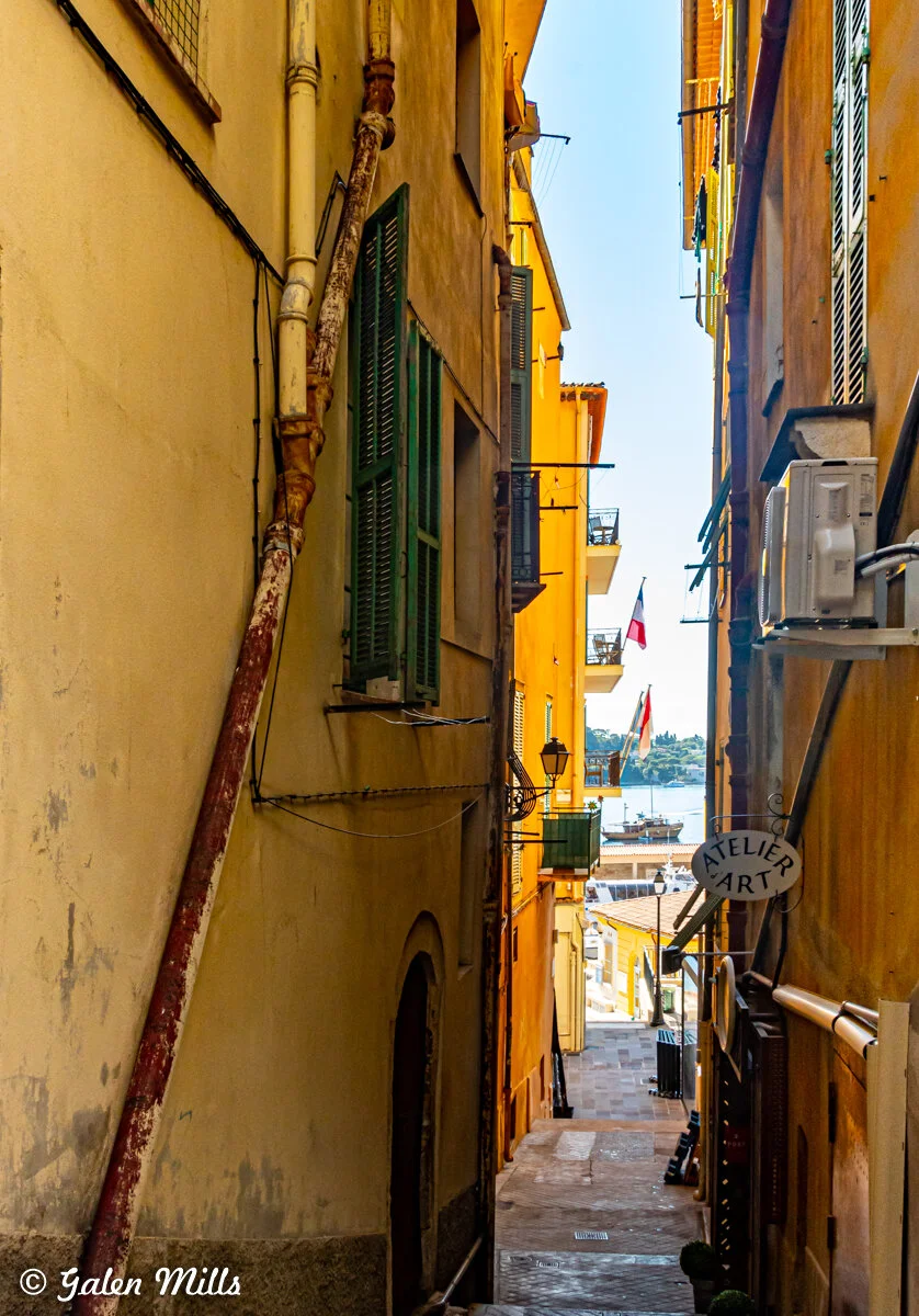 Narrow European alleyway with colorful buildings and a view of the sea in the background. Flags and an "Atelier Art" sign are visible.