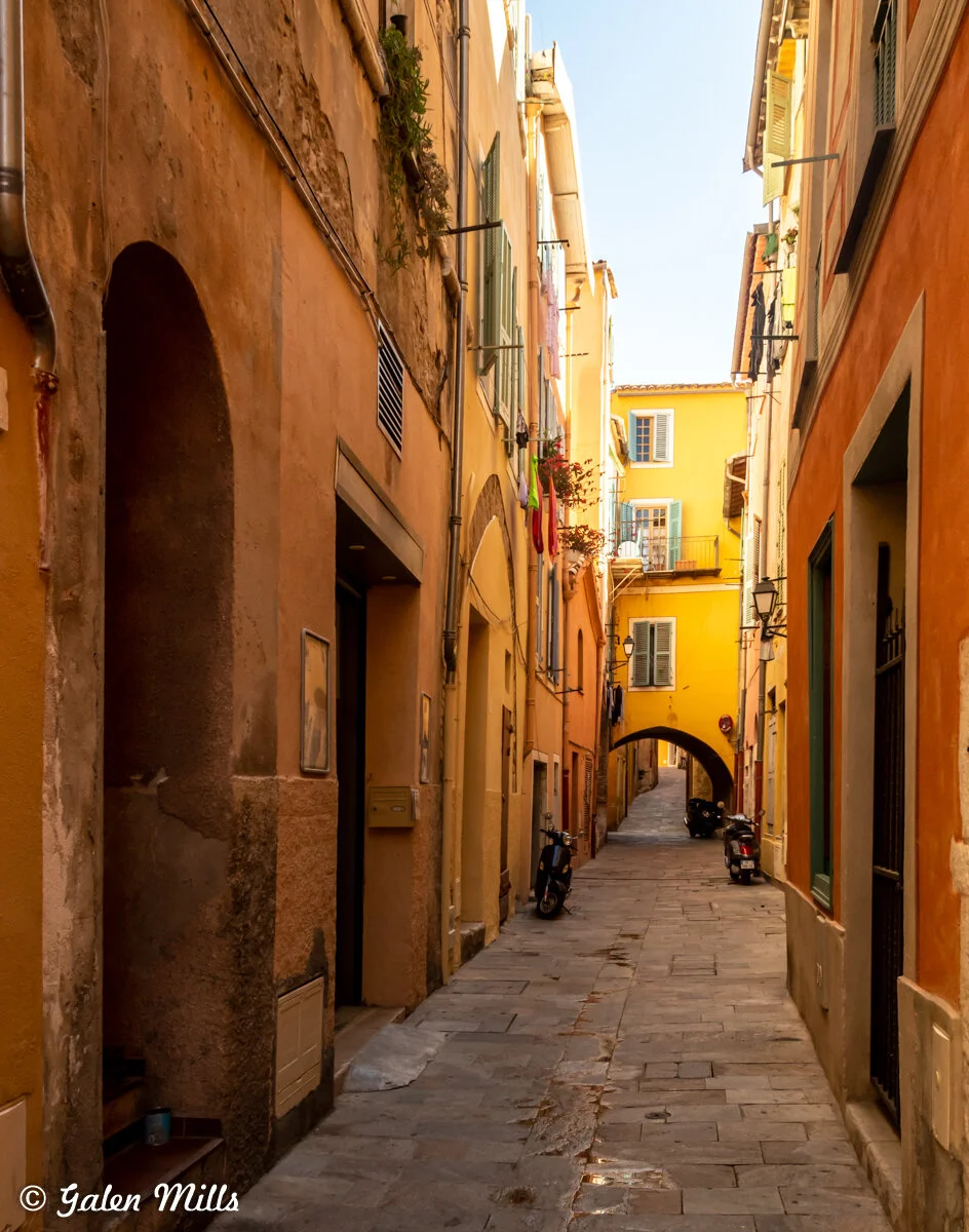 Narrow alleyway in a European town with stone pavement, colorful buildings, and motorbikes. An archway is visible in the background.