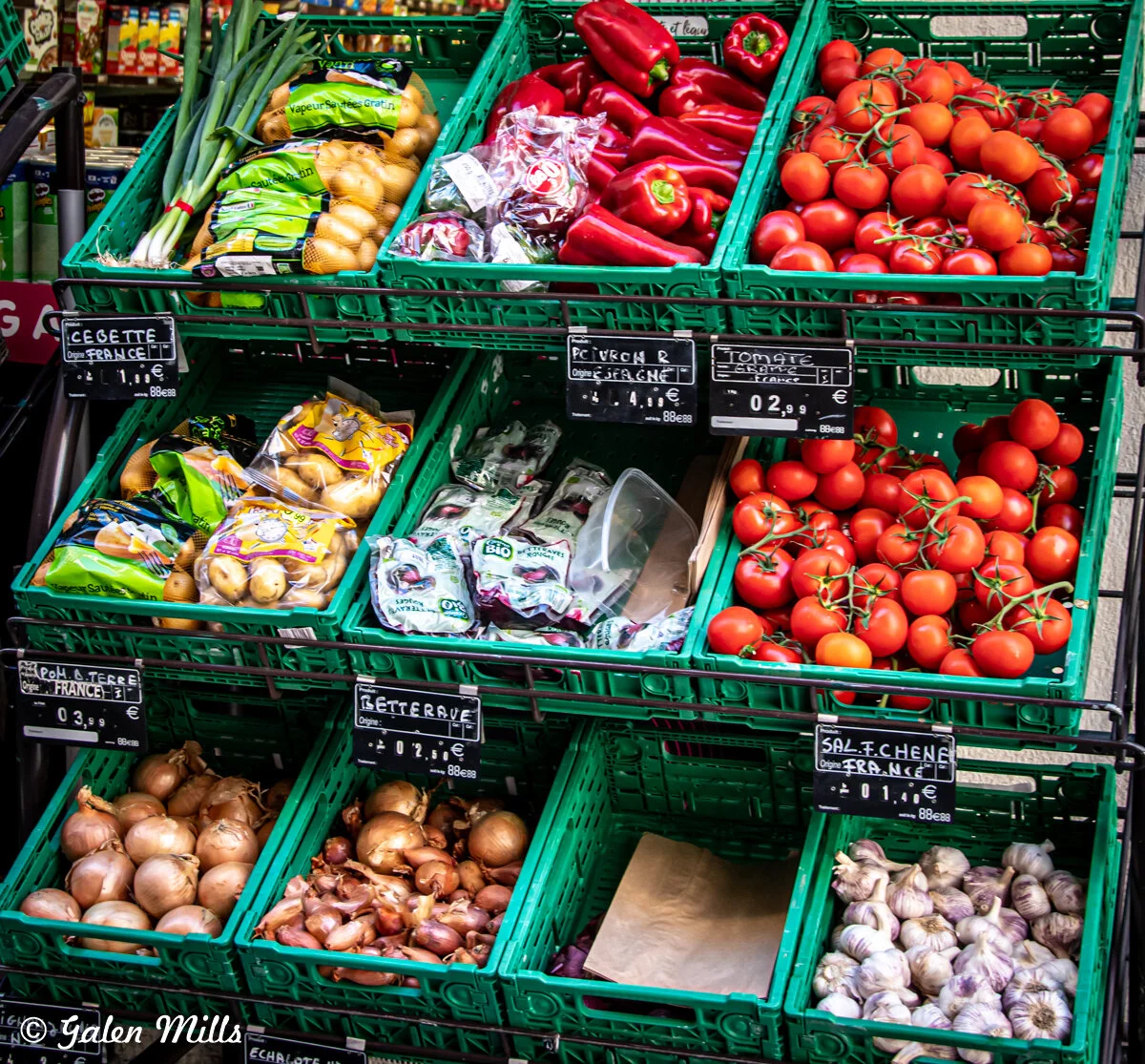 Display of fresh vegetables including onions, garlic, tomatoes, red peppers, potatoes, and green onions in green plastic crates at a market, with price tags in French.