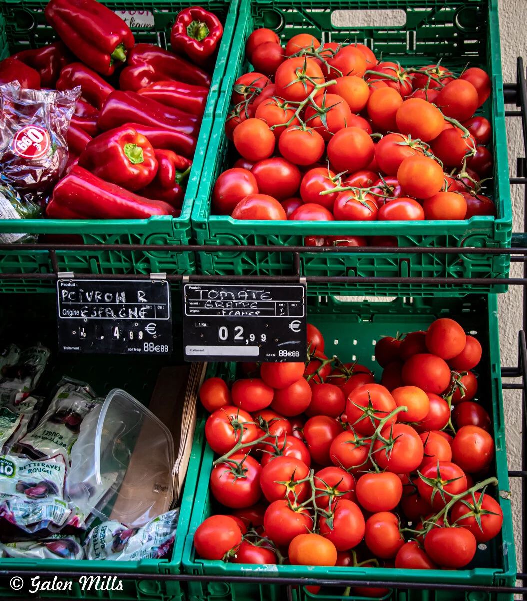 Crates of red bell peppers and vine tomatoes with price tags in a market display.