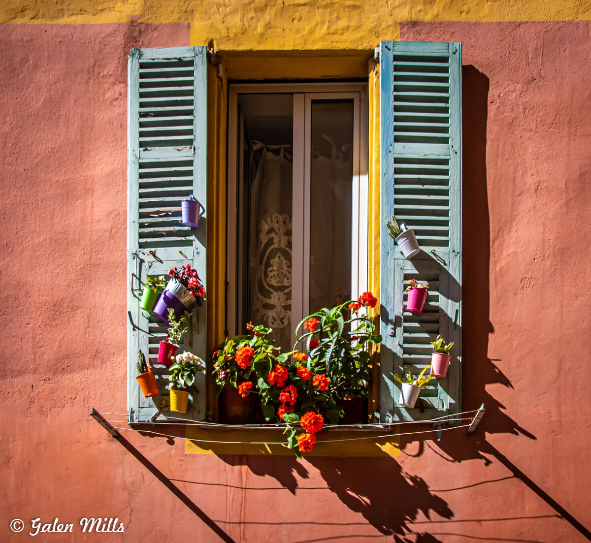 Colorful window with blue shutters, red flowers, and hanging small pots on a vibrant wall.