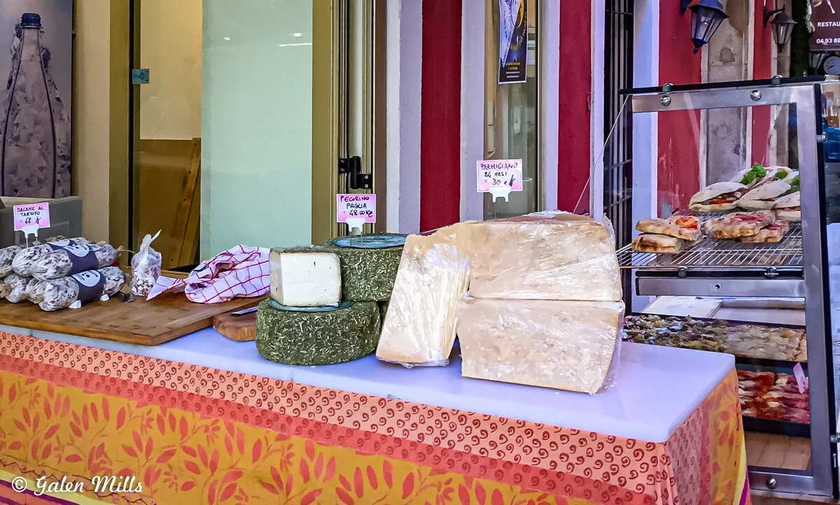 Cheese and salami on display at an outdoor market stall, including large wedges of Parmigiano cheese and a selection of salami packed on a wooden board, with sandwiches in a warmer.