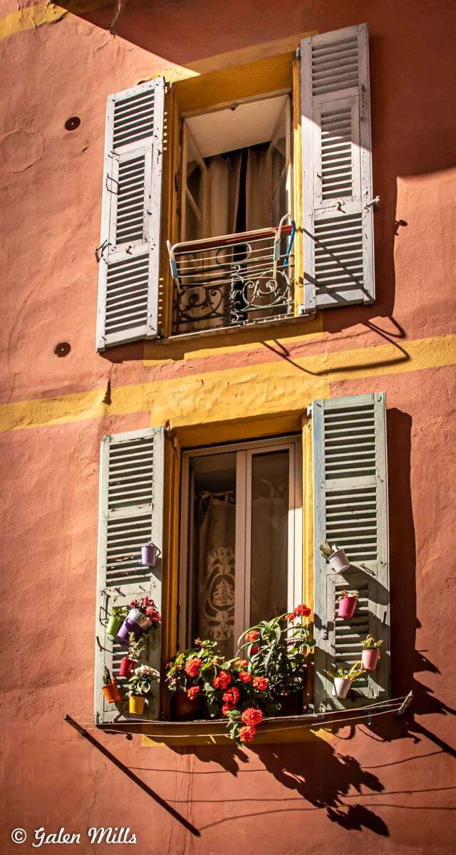 Two windows with blue shutters on a peach-colored wall, adorned with flower pots and red flowers. Sunlight casts shadows on the wall.