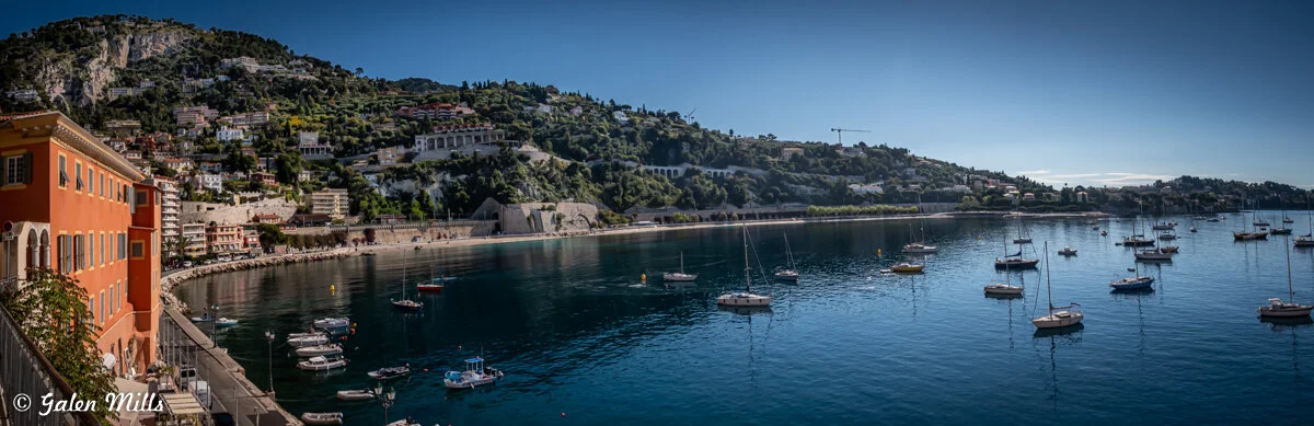 Panoramic view of a coastal town with boats on calm water, lush green hills, and colorful buildings along the shoreline under a clear blue sky.