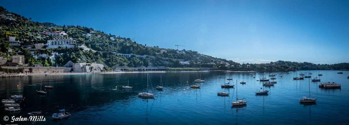 Panoramic view of a bay with several anchored boats, surrounded by hills with lush greenery and buildings under a clear blue sky.