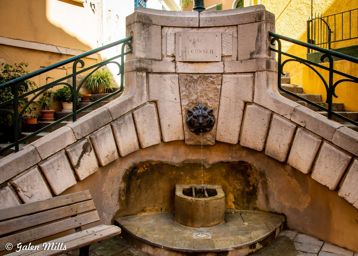 Stone fountain with lion head spout in historic setting, flanked by stairs and potted plants, labeled "Place du Conseil."