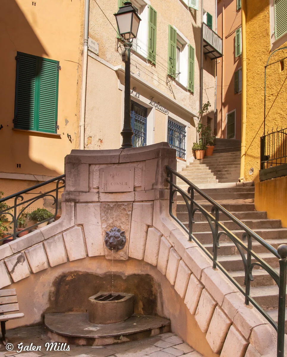 Stone fountain with lion's head spout, surrounded by buildings with colorful shutters, a street lamp, and stairs in a European street scene.