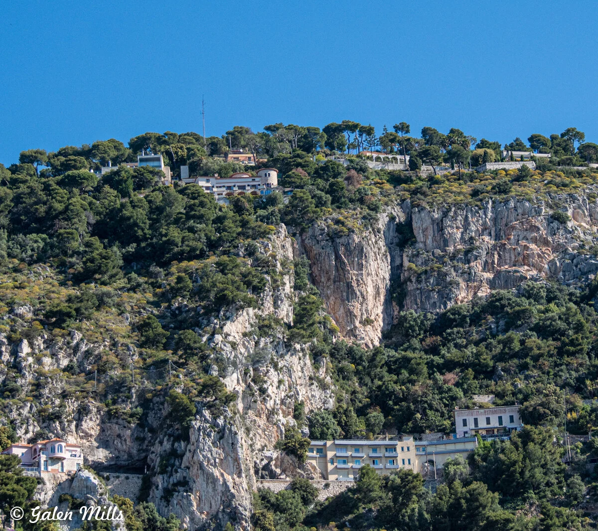 Scenic view of Mediterranean coastline with steep rocky cliffs and green vegetation, featuring buildings atop and along the cliffs.