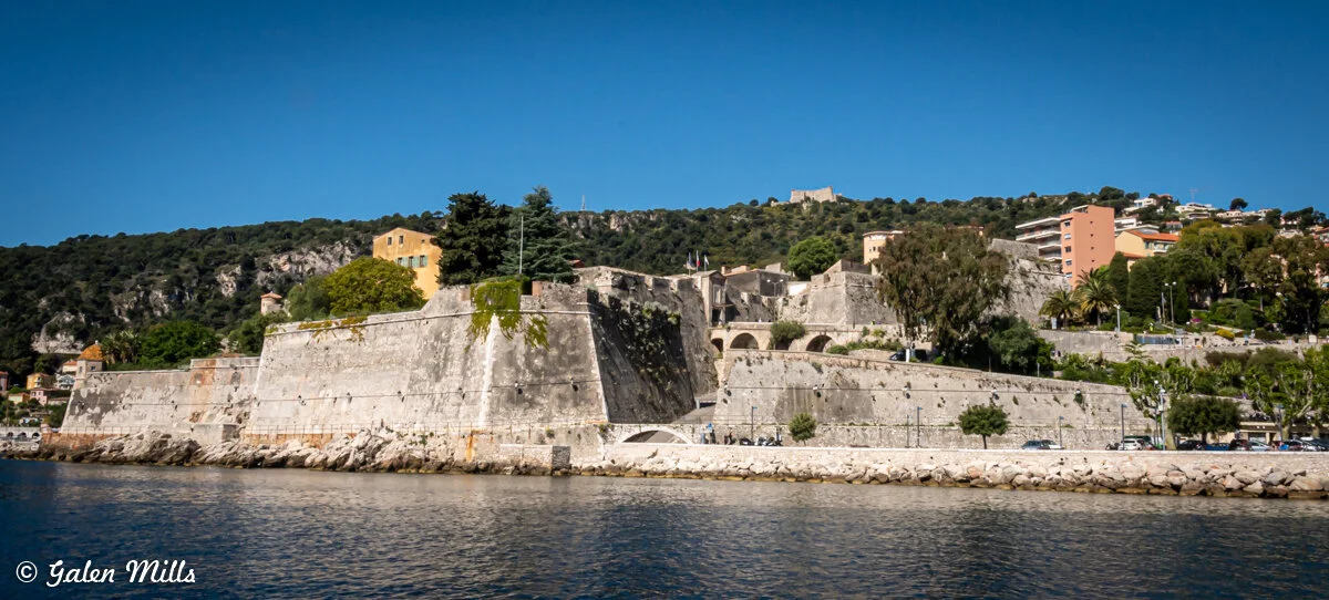 Fortress on a hillside by the water under a clear blue sky, with trees and buildings around.