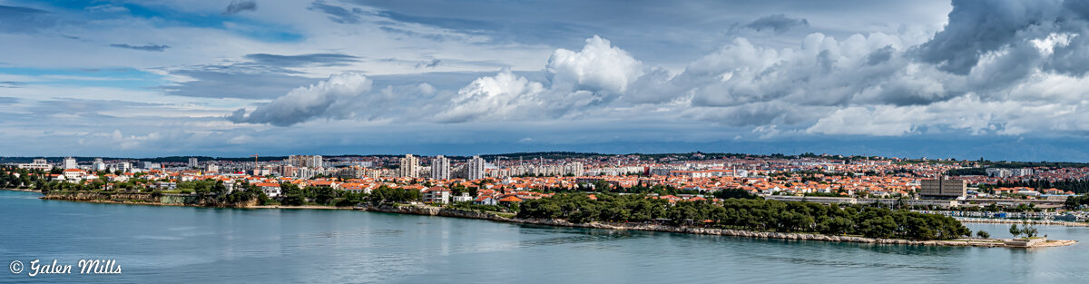 Panoramic view of Zadar, Croatia, featuring a coastline with greenery, historical buildings, and modern apartments under a partly cloudy sky.