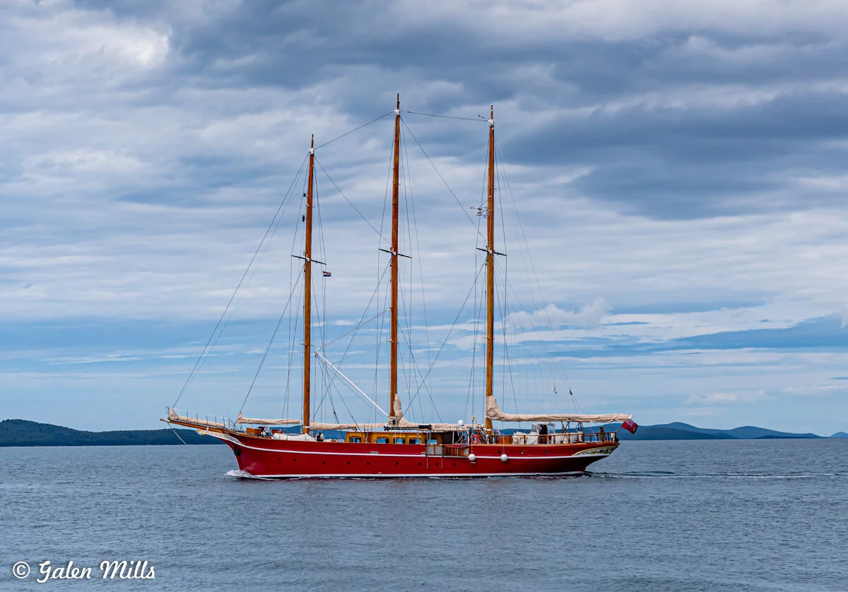 A large red sailboat with three tall masts sailing on calm water under a cloudy sky.
