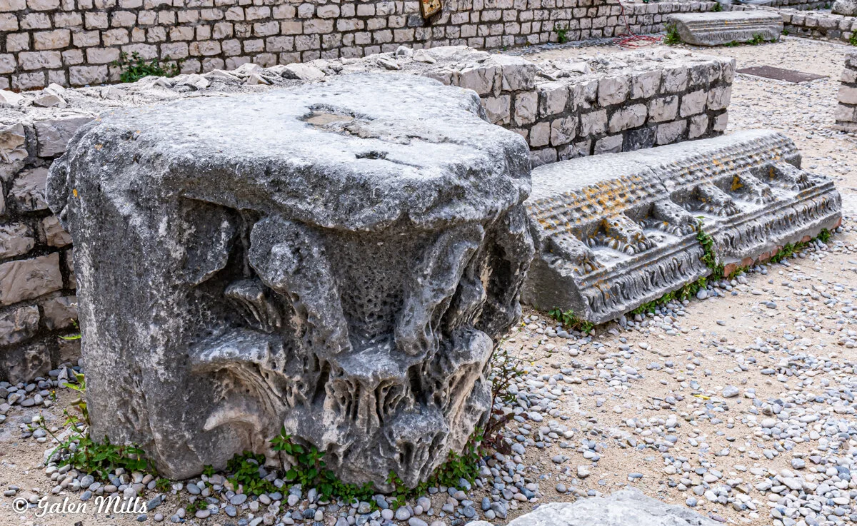 Ancient stone ruins with carved details on a cobblestone surface, surrounded by smaller stone blocks and walls.