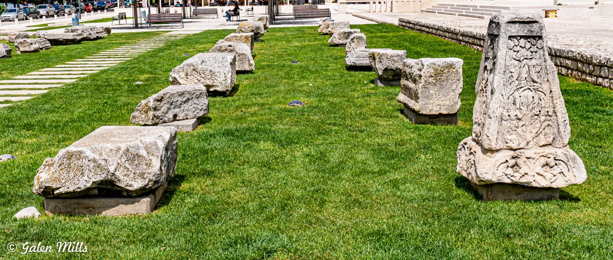 Ancient stone artifacts arranged on grass outdoors, with one engraved stone in the foreground, in a park-like area.