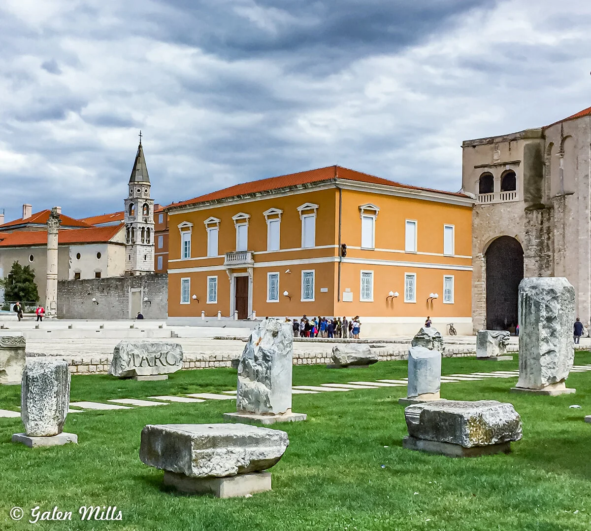 Historical site with ancient stone ruins in foreground, yellow building with red roof in center, bell tower in background, cloudy sky.