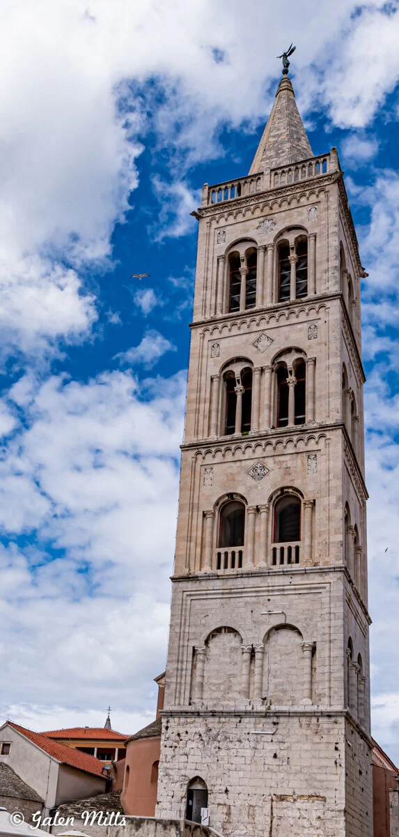 Tall stone bell tower with arched windows under a blue sky with clouds.