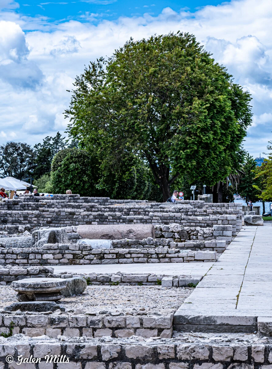 Ancient stone ruins with people sitting nearby, large leafy tree in background, cloudy sky above.