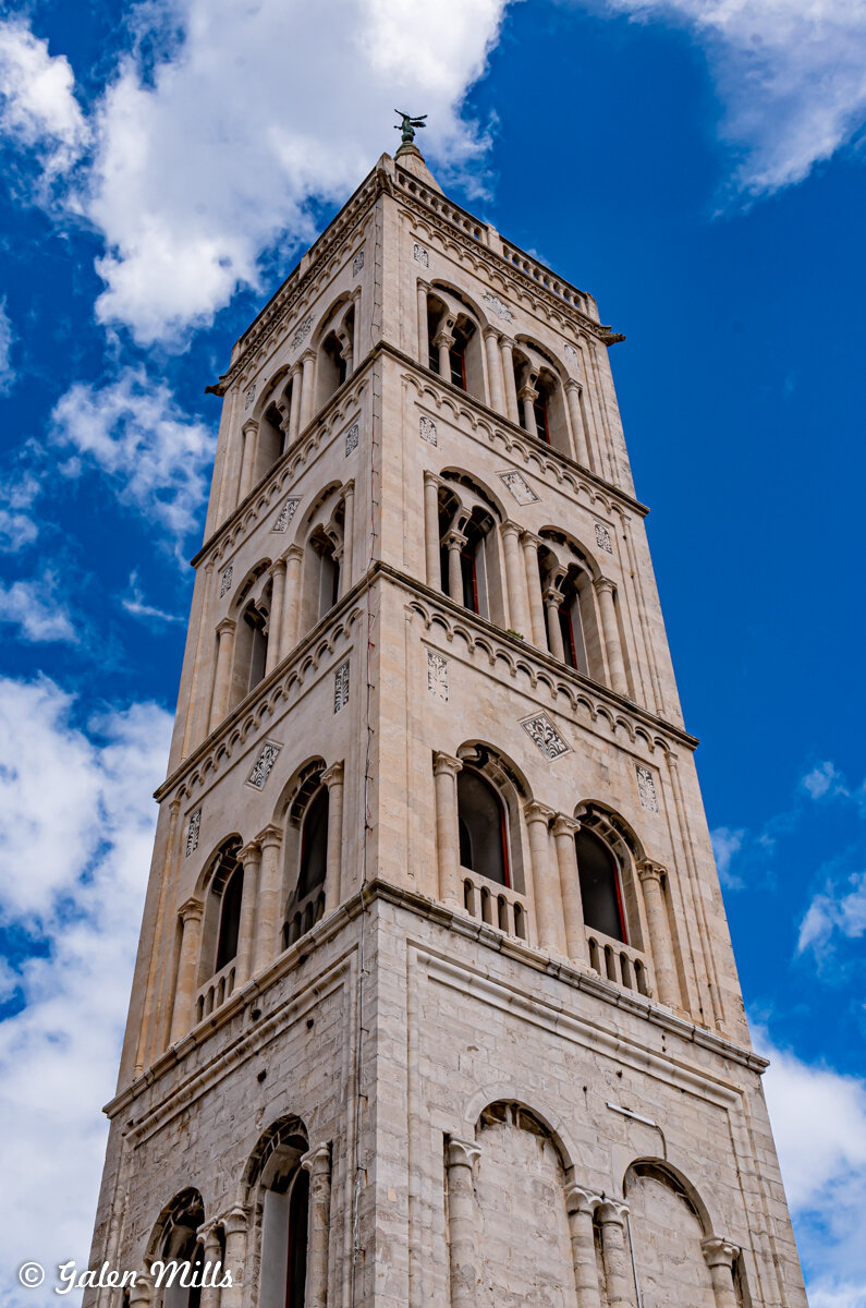 Stone bell tower with arched windows against a blue sky with clouds.