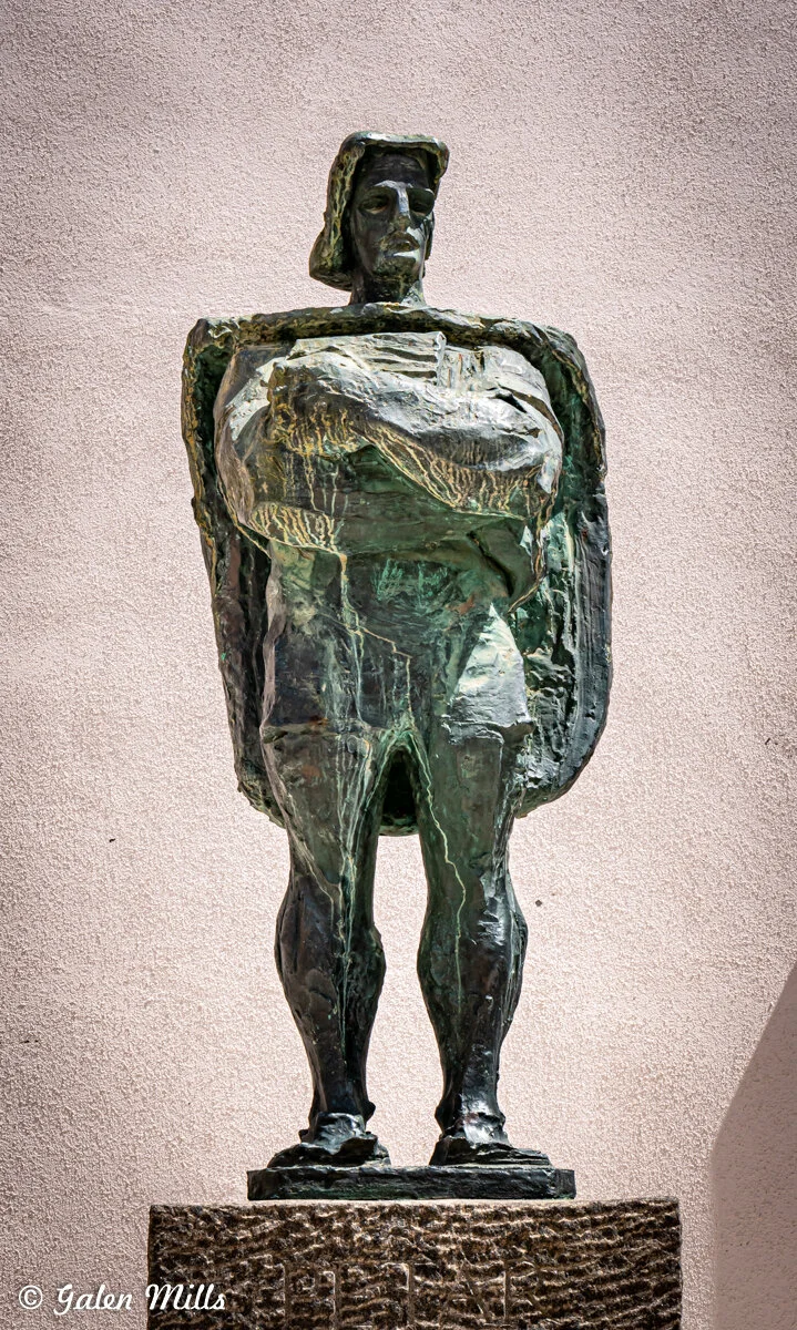 Bronze statue of a standing figure with arms crossed, on a textured stone pedestal against a plain background.