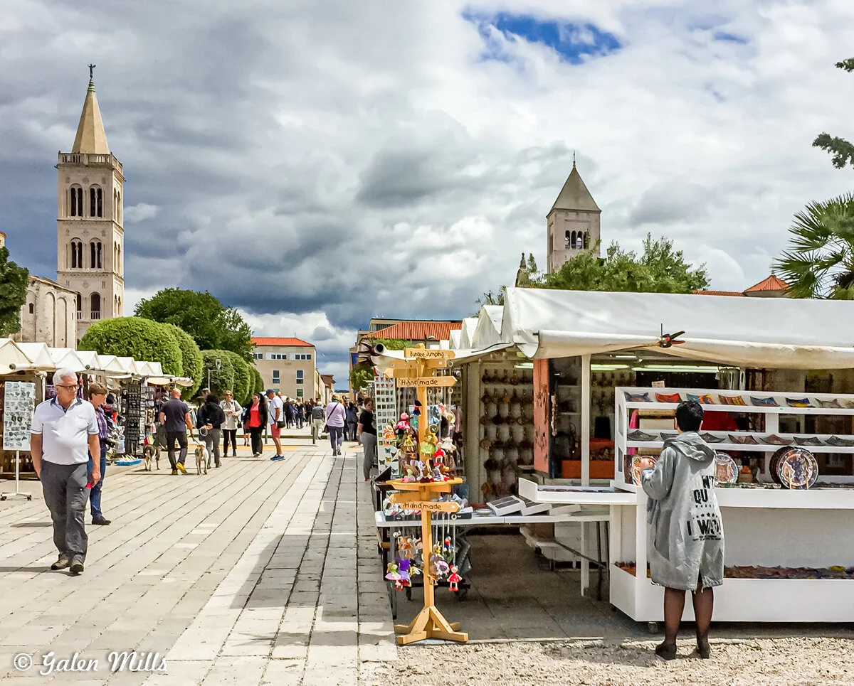 Outdoor market scene with stalls selling handmade crafts, with people walking and shopping, and a church tower visible in the background.