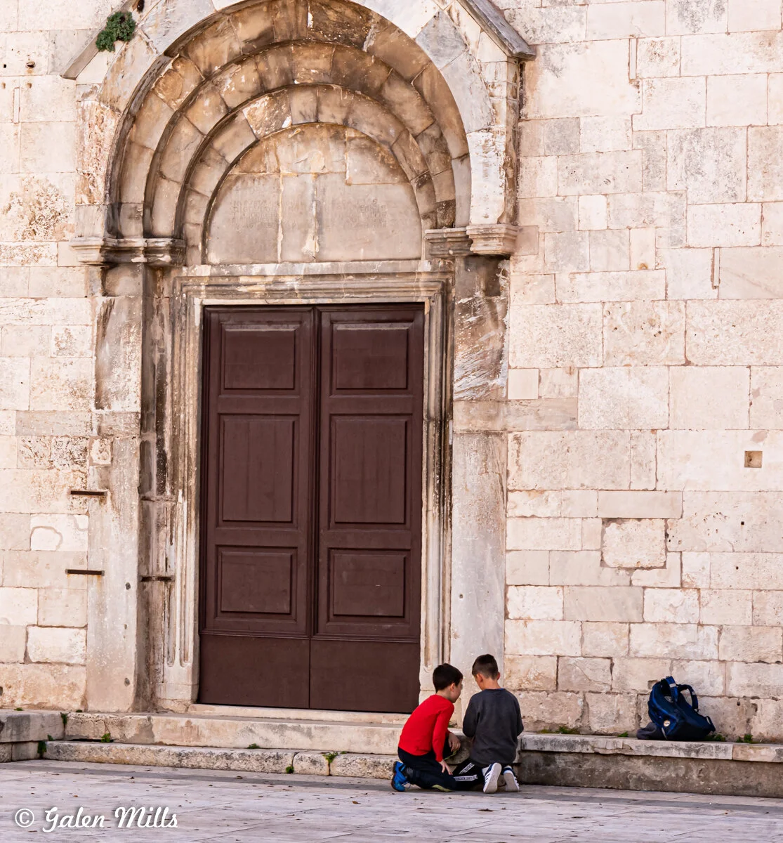 Two boys sitting on a stone ledge outside a historic stone building with a large brown wooden door, engaged in conversation or play, with a backpack nearby.