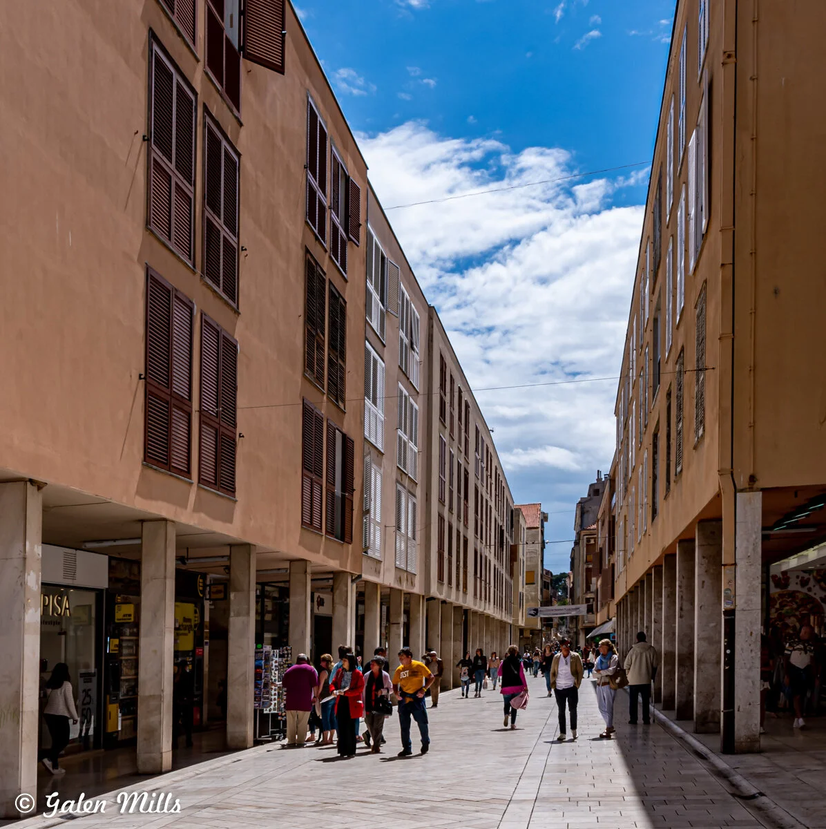 Pedestrian street in a modern urban area lined with buildings featuring large windows and shutters, with people walking under a partly cloudy sky.