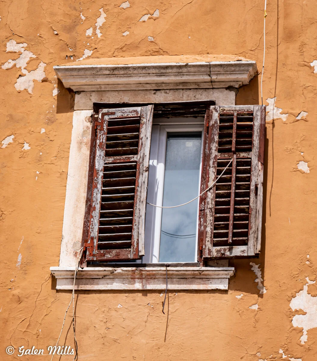 Old window with peeling paint and open wooden shutters on a weathered wall.