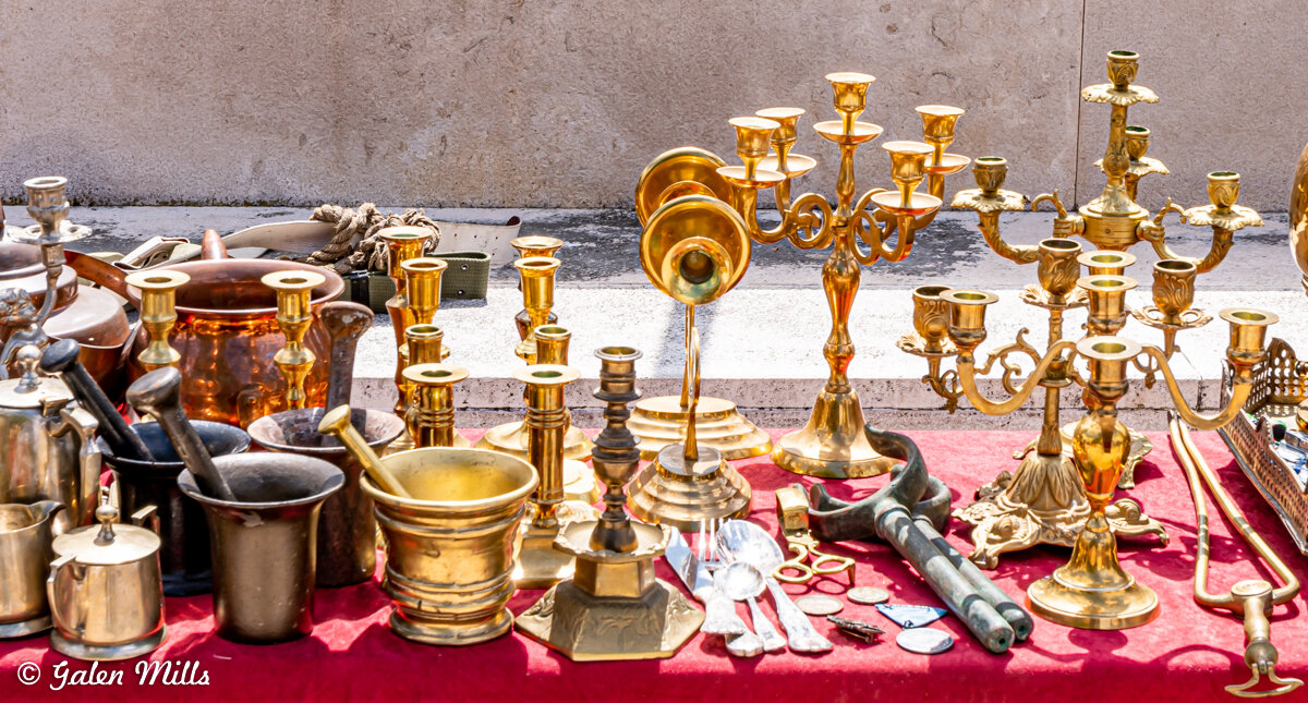 A variety of vintage brass and metal items, including candlesticks, mortars, pestles, and other decorative objects, displayed on a red cloth at a flea market or antique shop.