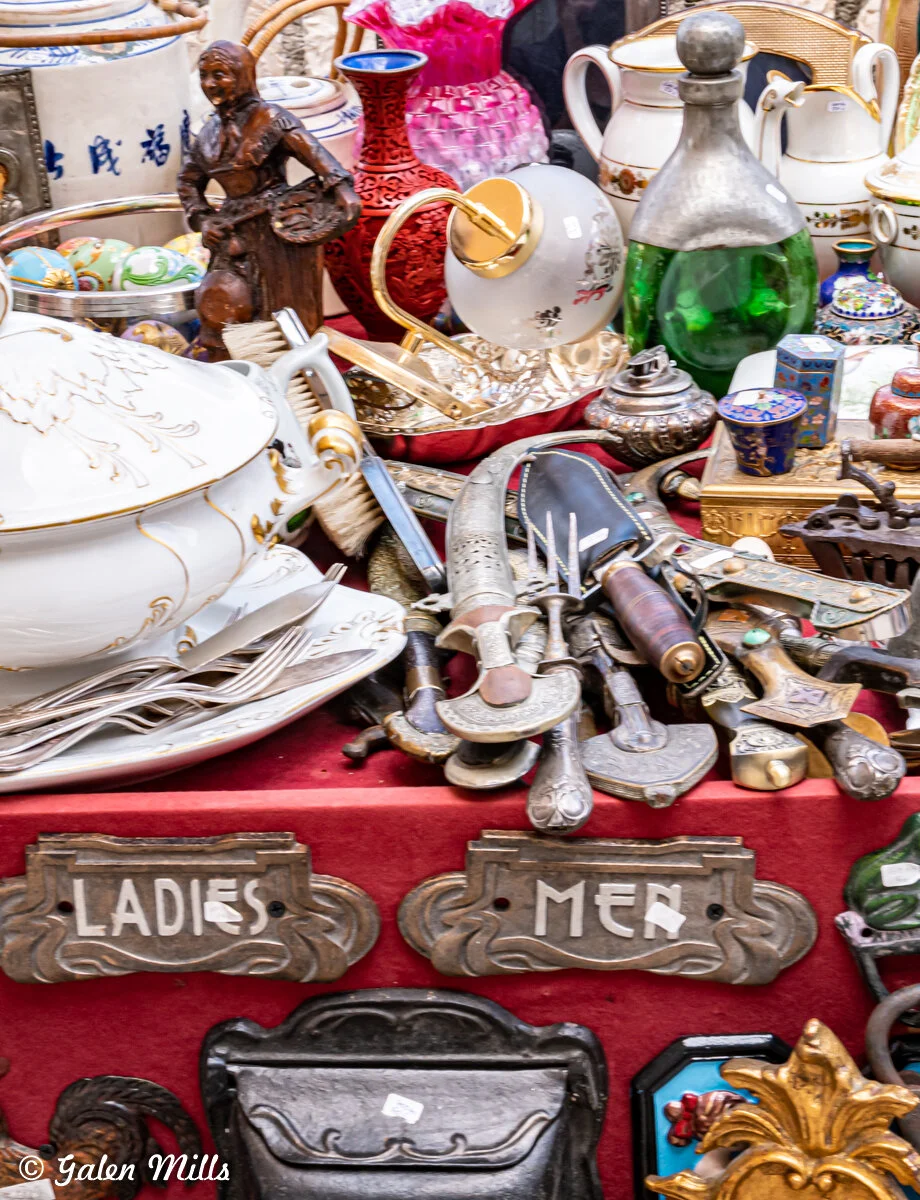 Antique and vintage items on display at a market, including ornate dishes, teapots, knives, and decorative signs labeled 'Ladies' and 'Men'. Various colorful vases and a figurine are also visible.