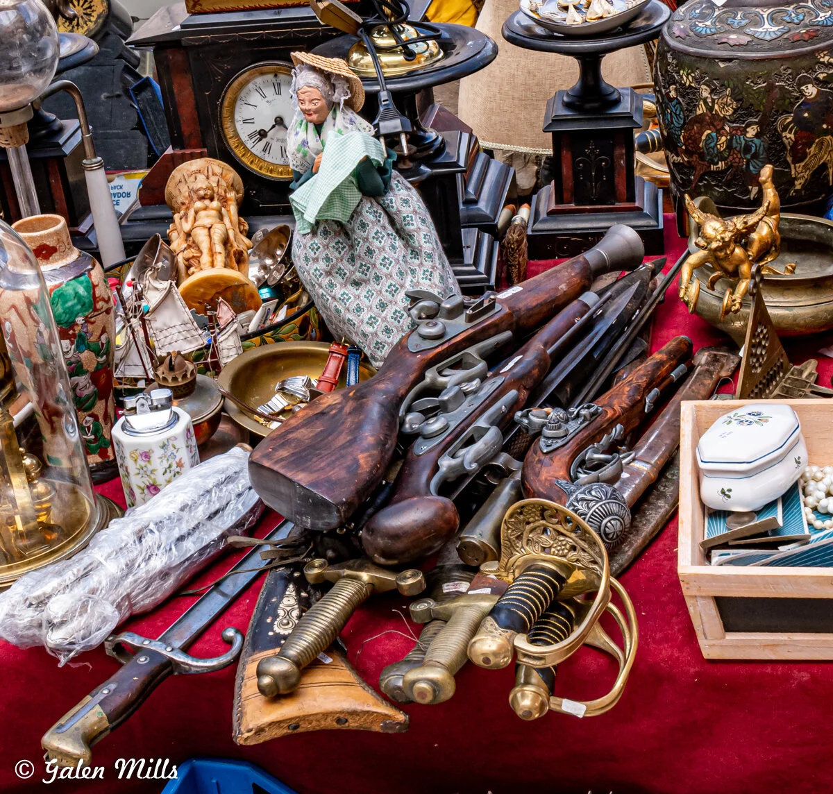 A collection of antique items displayed on a table, including old rifles, pistols, swords, a ceramic doll, a clock, a lantern, a decorative box, and various small figurines and trinkets.