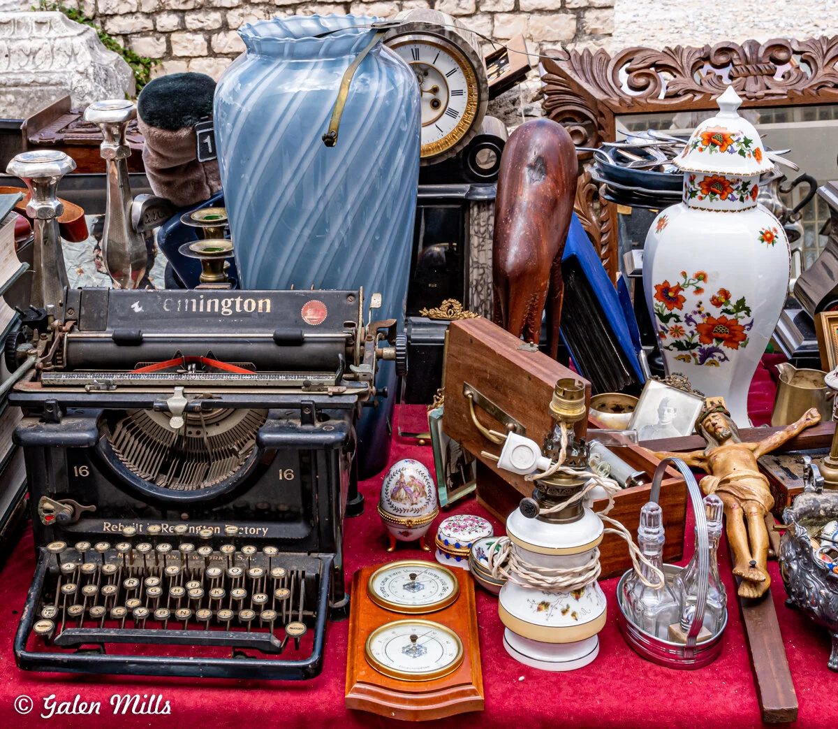 Antique market display featuring a vintage typewriter, large blue vase, clock, decorative porcelain jar, religious figurine, assorted trinkets, and miscellaneous collectibles on a red tablecloth.