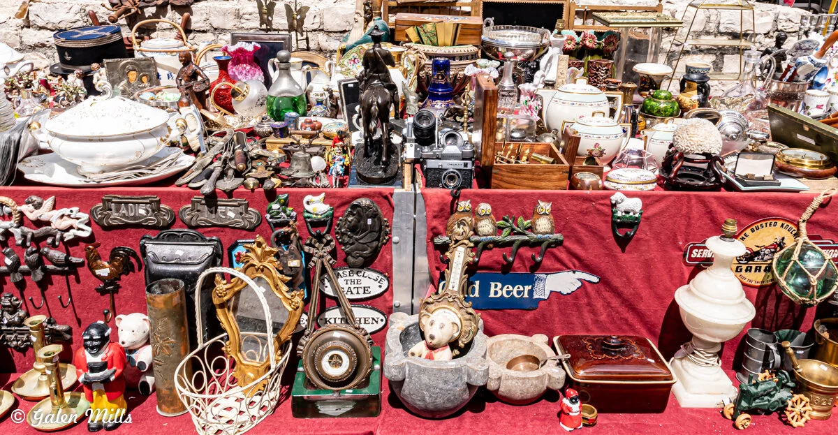 A flea market table filled with vintage items, including ceramic dishes, decorative owls, old cameras, figurines, and various metal and wooden knick-knacks, all displayed on a red tablecloth.