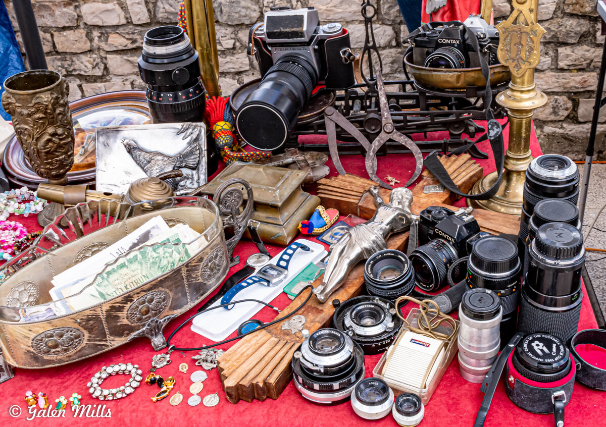 A collection of various vintage items displayed on a red tablecloth, including old cameras, lenses, a brass candlestick, a carved metal box with money, watches, a silver figurine, a bronze cup, antique scissors, and decorative plate. The background f