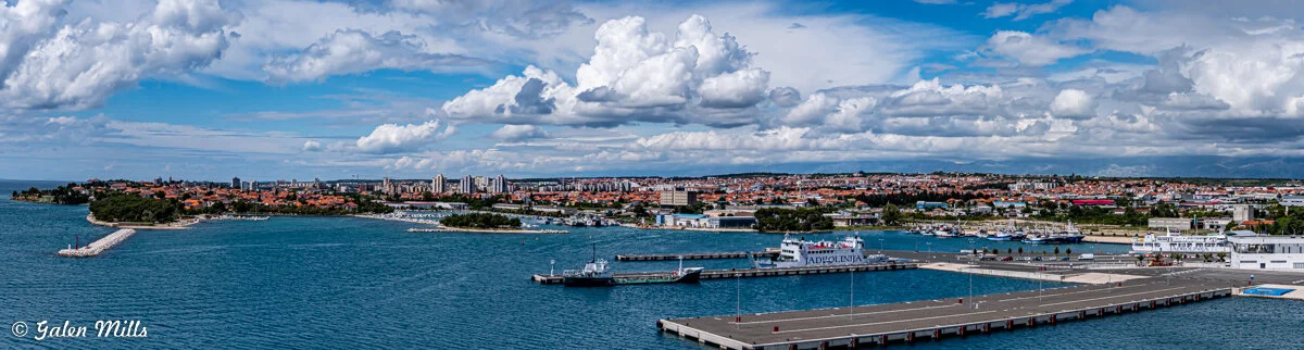 Panoramic view of a coastal city with waterfront, harbor, docked boats, and city skyline under a cloudy sky.