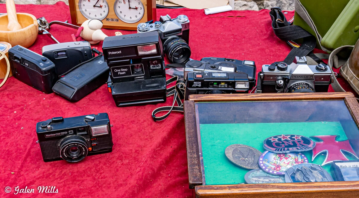 Various vintage cameras and equipment displayed on a red cloth, including a Polaroid One Step Flash, a Ricoh camera, and other film cameras. A clock and a glass display case with pins or patches are also visible.