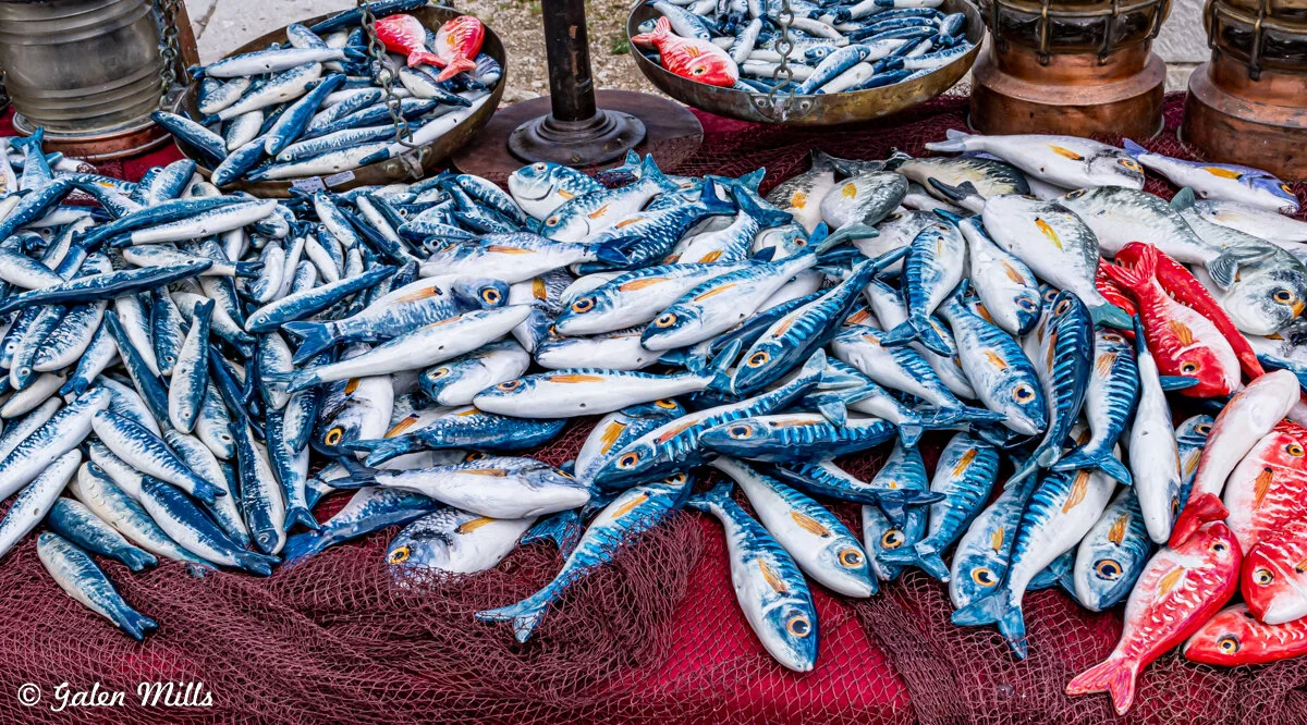 Pile of colorful ceramic fish with blue and red detailing on a net, displayed with metal lanterns.