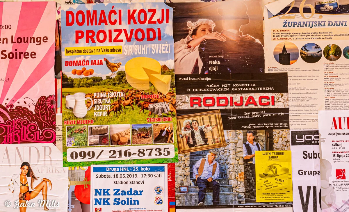 A colorful display of various posters, including advertisements for local goat products, theatrical performances, and events. Prominent text in Croatian includes offers for cheese, yogurt, and kefir, alongside promotional material for plays and commu
