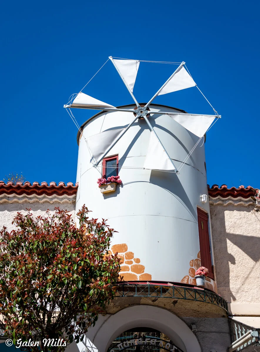 Windmill structure with white blades on a white cylindrical building, red-framed window with pink flowers, tree in foreground, under a clear blue sky.